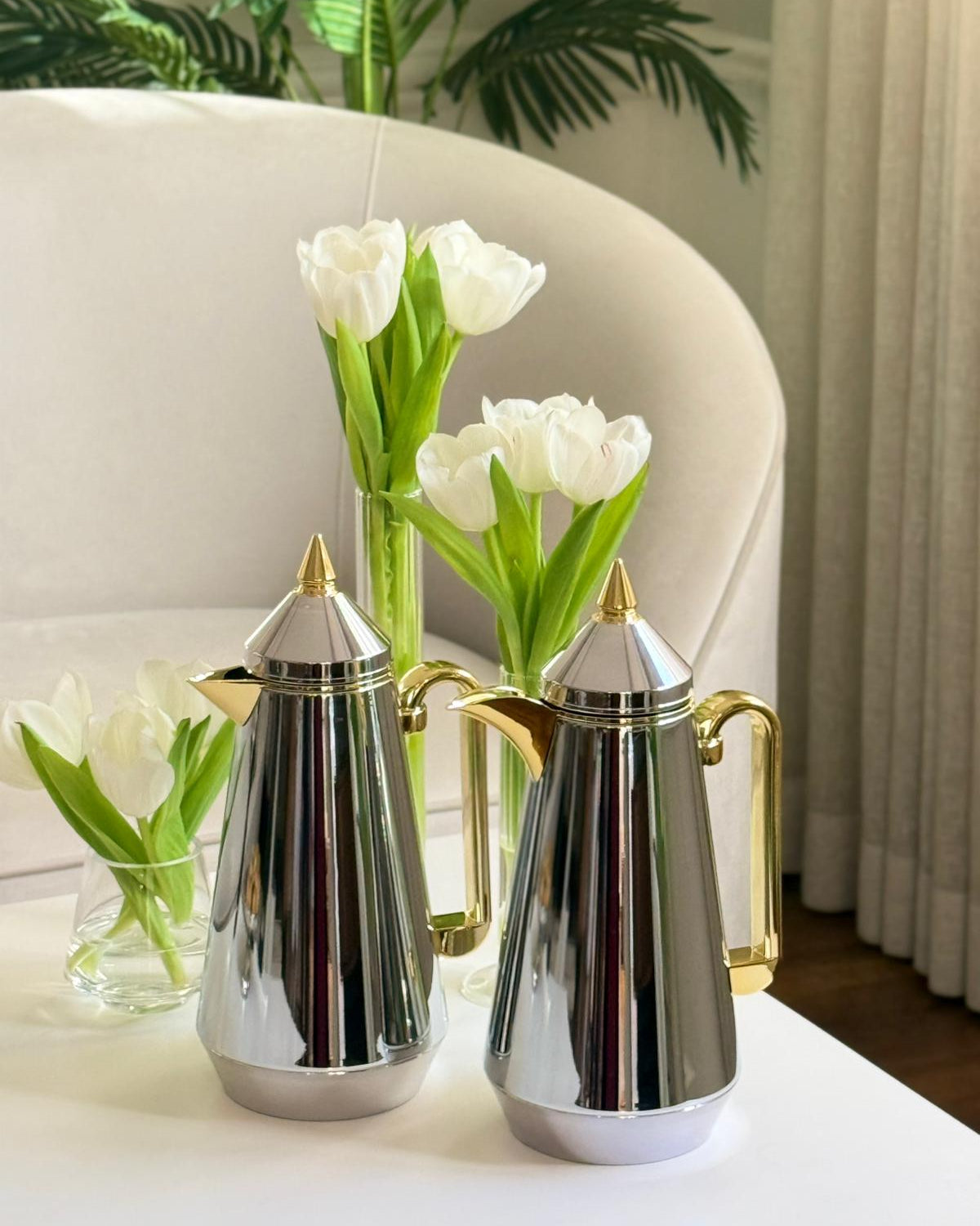 Two silver and gold coffee pots on a table with white tulips in the background.