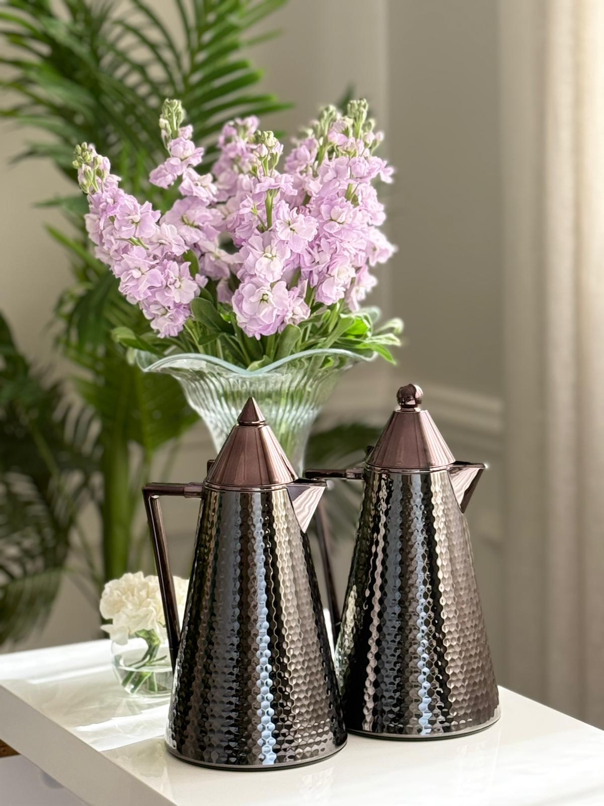 Two metallic tea pots on a table with a vase of pink flowers in the background.