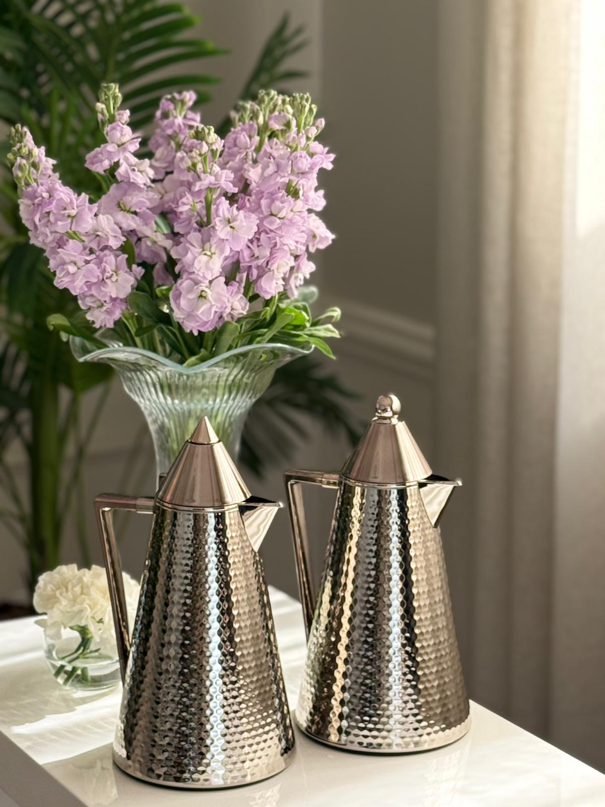 Two metallic teapots on a table with a vase of purple flowers in the background.